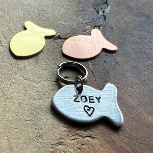 May include: A silver fish-shaped pet tag with the name "ZOEY" engraved on it, along with a small heart. The tag is attached to a silver key ring. There are two other fish-shaped tags in the background, one gold and one copper.