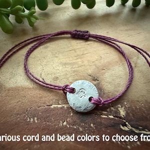 May include: A handmade bracelet with a silver-colored, hammered metal disc charm. The charm has an embossed design and is strung on a deep purple cord. The text "Various cord and bead colors to choose from" is at the bottom.