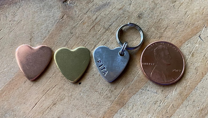 Pu&ograve; includere: Tre etichette di metallo a forma di cuore, una di rame, una di ottone e una d'argento con il nome "PITA" inciso su di essa. L'etichetta d'argento ha un portachiavi attaccato. Le etichette sono disposte su una superficie di legno accanto a un centesimo americano.