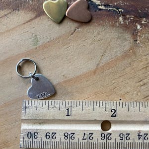 Pu&ograve; includere: Un'etichetta a forma di cuore argentata con la parola "PITA" incisa su di essa. L'etichetta &egrave; attaccata a un anello argentato e si trova accanto a un righello. Il righello &egrave; contrassegnato con pollici e frazioni di pollice.