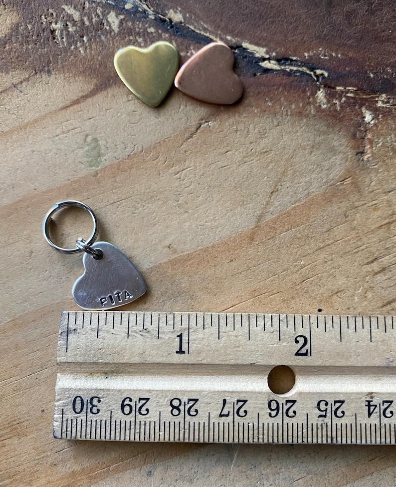 Pu&ograve; includere: Un'etichetta a forma di cuore argentata con la parola "PITA" incisa su di essa. L'etichetta &egrave; attaccata a un anello argentato e si trova accanto a un righello. Il righello &egrave; contrassegnato con pollici e frazioni di pollice.