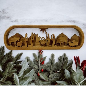 May include: A wooden cutout of the nativity scene with a star above the manger. The scene is framed in a wooden oval and features figures of Mary, Joseph, baby Jesus, a donkey, a sheep, and three wise men.