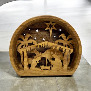 May include: A wooden nativity scene, featuring a layered design with a star, palm trees, and silhouettes of figures. The piece is crafted from light brown wood, with a semi-circular shape and a flat base. The design includes stars and a central star.