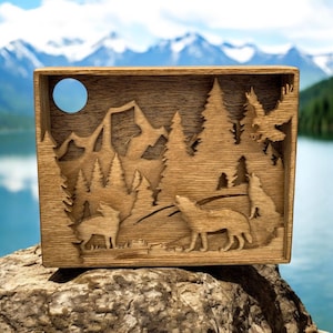 May include: A wooden wall art piece with a carved design of a forest scene with mountains, trees, and wolves. The piece is painted in a dark brown color and has a rustic look.