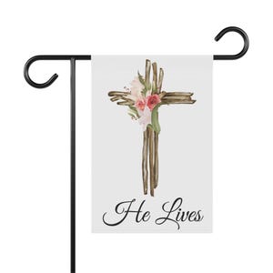 Könnte beinhalten: Eine weiße Gartenflagge mit einem braunen Holzkreuz, das mit rosa und weißen Blumen verziert ist. Der Text "He Lives" ist in schwarzer Kursivschrift unter dem Kreuz geschrieben.