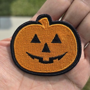 May include: A small orange and black embroidered patch of a jack-o'-lantern with a toothy grin.