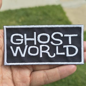 Può includere: Una toppa rettangolare nera con testo ricamato bianco che recita "GHOST WORLD". La toppa ha un bordo bianco. La toppa è tenuta in una mano su uno sfondo di erba verde.