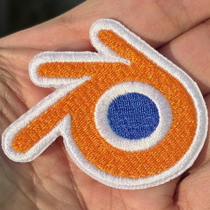 Pode incluir: Patch bordado laranja e branco com um círculo azul no centro, que se assemelha ao logotipo do software Blender 3D.