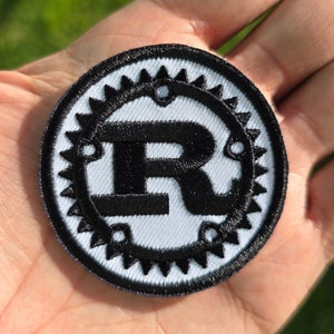May include: A black and white embroidered patch with a stylized letter "R" in a circular gear design.