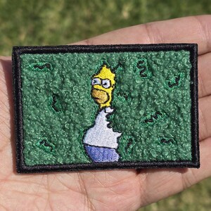 May include: Embroidered patch featuring a cartoon character with yellow skin, wearing glasses, a white shirt, and blue pants. The patch has a green textured background with black border.