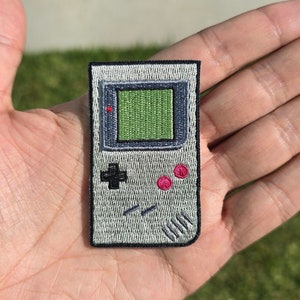 May include: A gray and black embroidered patch of a handheld video game console. The console has a green screen, a black directional pad, and two red buttons.