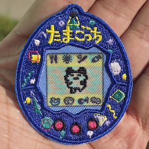 以下が含まれることがあります： 青い刺繍パッチ、たまごっちの形。パッチには、黄色の日本語の「たまごっち」、ピクセル化されたキャラクター、さまざまな色の装飾要素が描かれています。