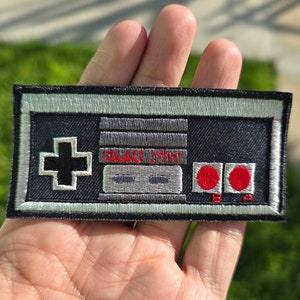 May include: A black and silver embroidered patch shaped like a vintage video game controller with the text "BLACK HISTORY" in red. The patch has a red plus sign, two gray buttons, and two red circles.