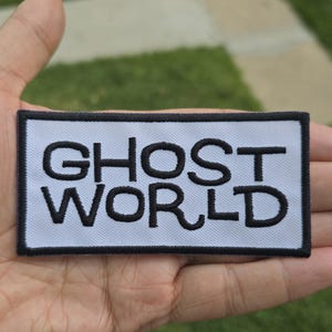 Può includere: Una toppa rettangolare con le parole "GHOST WORLD" ricamate in nero su sfondo bianco. La toppa ha un bordo nero. La toppa è tenuta in mano su uno sfondo di erba verde.