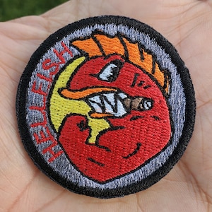 Peut inclure: Un patch rond brodé de couleur gris et noir avec un poisson de dessin animé rouge, jaune et orange tenant une cigarette dans sa bouche. Le texte "YELLFISH" est brodé autour du poisson avec un fil blanc.