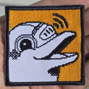 Könnte beinhalten: Ein weißer gestickter Aufnäher mit einem Cartoon-Bild eines weißen Roboters mit einem schwarzen Auge und einem schwarzen Mund, der eine Schallwelle erzeugt. Der Roboter befindet sich in einem gelben Quadrat mit schwarzem Umriss.