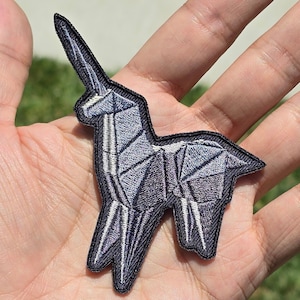 May include: A black and white embroidered patch of an origami unicorn. The unicorn is standing on all four legs with its horn pointing upwards.