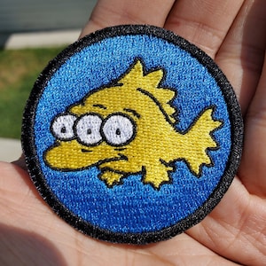 May include: An embroidered patch featuring a cartoon fish with three eyes. The fish is yellow and is set against a blue background with a black border.