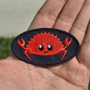 May include: A red embroidered crab with white eyes on a black oval patch. The crab is smiling and has two claws.