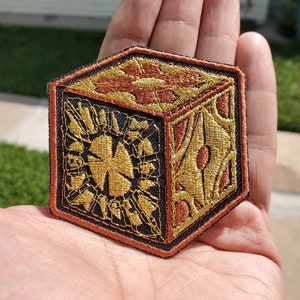 Könnte beinhalten: Ein brauner und goldener gestickter Patch in Form eines Würfels. Der Würfel hat ein detailliertes Design mit komplizierten Mustern und einem goldenen Stern in der Mitte.