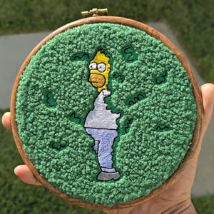 May include: Embroidered artwork in a wooden hoop, depicting Homer Simpson from The Simpsons. The character is in yellow, white, and blue, set against a textured green background. The artwork is held in a hand, with the wooden hoop visible.