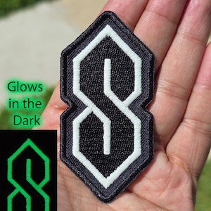 May include: A black embroidered patch featuring the iconic geometric S shape outlined in white thread. The patch includes a secondary inset showing the design in a glowing green state with the text Glows in the Dark printed next to it.
