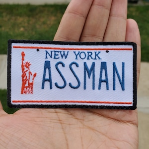 Könnte beinhalten: Ein weißer Patch mit einem blauen und roten gestickten Bild der Freiheitsstatue und dem Text "NEW YORK ASSMAN" in blauen und roten Buchstaben. Der Patch hat einen schwarzen Rand und einen roten und orangenen Rand.