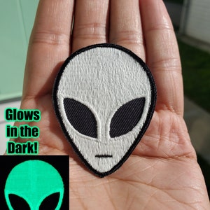 May include: White alien face embroidered patch with black eyes and mouth. The patch glows in the dark and is shown on a hand. The glow in the dark version of the patch is shown in a smaller image next to the hand.