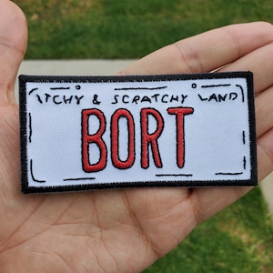 May include: A white embroidered patch with black outlines. The patch has the text "ITCHY & SCRATCHY LAND" at the top and "BORT" in red letters in the centre.