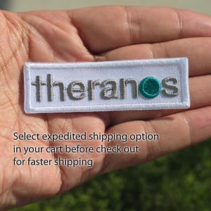 May include: A rectangular white embroidered patch featuring the word theranos in grey lowercase letters, with a teal circle replacing the letter o. The patch has a white stitched border and is designed for application onto fabric surfaces.