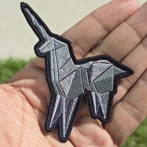 May include: An embroidered patch featuring a silver-colored unicorn designed with geometric, origami-style lines. The unicorn figure is outlined in a thick black border, with subtle purple shading visible within the lower sections of the legs.