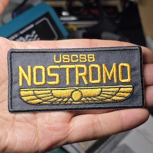 以下が含まれることがあります： 金色の文字で「USCSS NOSTROMO」と書かれた長方形のパッチ。その下に金色のエンブレムがあります。パッチは濃い灰色の背景にあります。