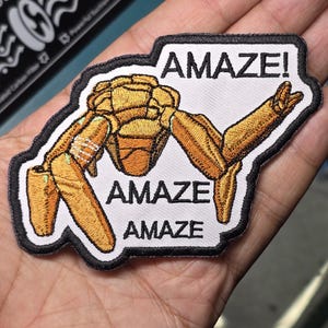 May include: Embroidered patch featuring a stylized, golden-yellow robot with the word "AMAZE!" above and "AMAZE" repeated below. The patch has a black border and is on a white background.