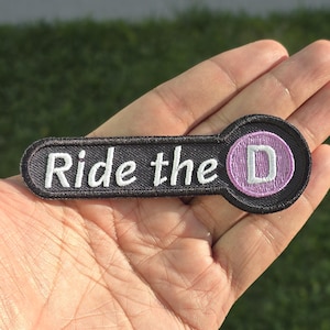 May include: Black and purple embroidered patch with the text "Ride the D". The patch has a circular design with the letter "D" in white on a purple background. The patch is held in a person's hand.