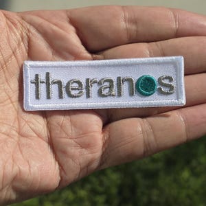 May include: A rectangular embroidered patch with a white background and a white stitched border. The text "theranos" is embroidered in grey thread, with the letter "o" replaced by a solid teal circular emblem.