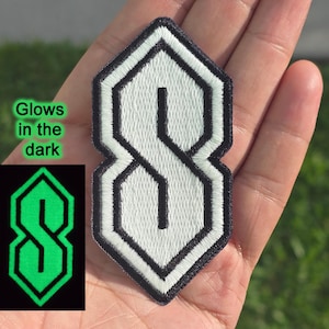 May include: Embroidered patch with the classic "S" design in white, outlined in black, and a distinctive geometric shape. A smaller image shows the design glowing green. Text on the left says "Glows in the dark".