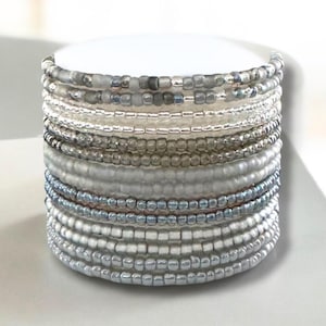 May include: A cylindrical beaded bracelet stack in shades of silver, gray, and clear. The stack features multiple rows of small, round beads, creating a textured and layered effect. The beads vary in size and color, adding depth and visual interest.