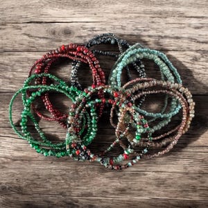 May include: A collection of beaded bracelets in various colors, including red, green, blue, and brown. The bracelets are made of small, round beads and are arranged on a wooden surface. The bracelets are a mix of solid colors and multicolored designs.