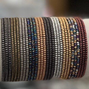 May include: A stack of 20 beaded bracelets in various colors, including silver, gold, gray, black, and red. The bracelets are made of small, round beads.