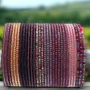 May include: A stack of beaded bracelets in shades of purple, pink, and orange. The bracelets are made of small, round beads and are arranged in a gradient pattern.