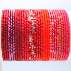 May include: A stack of red beaded bracelets with different textures and finishes. Some bracelets have a shiny iridescent finish, while others have a matte finish.