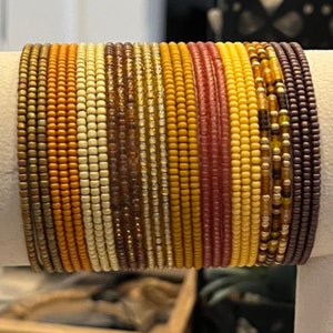 May include: A stack of colorful beaded bracelets in shades of brown, orange, yellow, and red. The bracelets are made of small, round beads and are arranged in a graduated pattern.