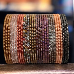 May include: A stack of beaded bracelets in various shades of copper, gold, and purple. The bracelets are made of small, round beads and are arranged in a vertical stack.