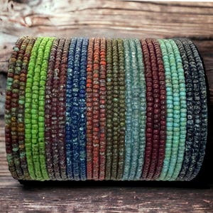 May include: A stack of colorful beaded bracelets in shades of green, blue, red, brown, and turquoise. The beads are small and uniform in size, creating a smooth and textured surface.