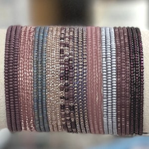 May include: A collection of beaded bracelets in various shades of purple, pink, and silver. The bracelets are tightly strung together, showcasing a range of colors from deep plum to pale rose. The beads appear to be small and uniform in size, creating a textured surface.