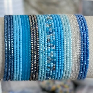 May include: A stack of blue and white beaded bracelets. The bracelets are made of small, round beads and are arranged in a gradient pattern, starting with dark blue and fading to white.