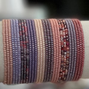 May include: A stack of beaded bracelets in shades of pink, purple, and gray. The bracelets are made of small, round beads and are arranged in a graduated pattern.