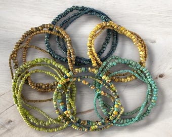 Puede incluir: Una colección de pulseras de cuentas en varios colores, incluyendo verde oliva, turquesa, amarillo mostaza y azul marino. Las pulseras están hechas de pequeñas cuentas redondas y están dispuestas en un patrón circular sobre una superficie de madera clara.