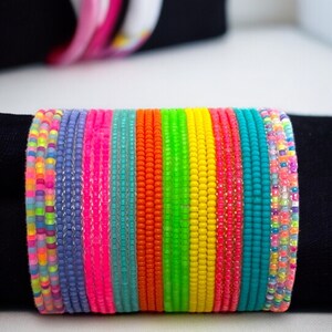 May include: A stack of colorful beaded bracelets in various neon shades, including pink, blue, green, yellow, and orange. The bracelets are made of small, round beads and are arranged in a rainbow pattern.