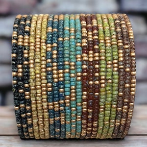 May include: A multi-colored beaded bracelet with gold accents. The bracelet is made of small, round beads in shades of blue, green, brown, and yellow. The beads are arranged in a striped pattern, with gold beads separating the different colors.